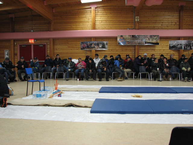 People seated in a semicircle in a wooden hall, with two parallel floor mats, an empty chair, and other supplies arranged in the centre.