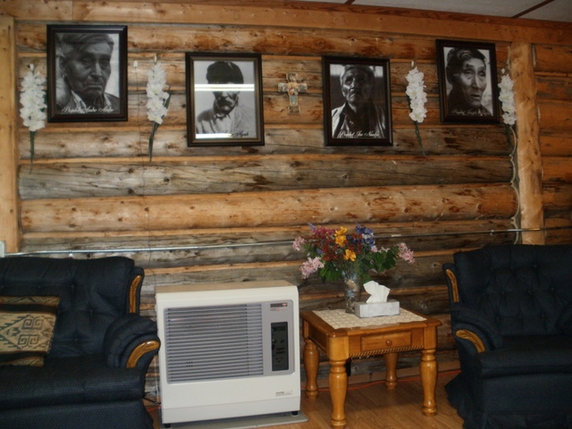Framed portraits of four Délı̨nę prophets displayed indoors on a log wall above chairs and a table with flowers.