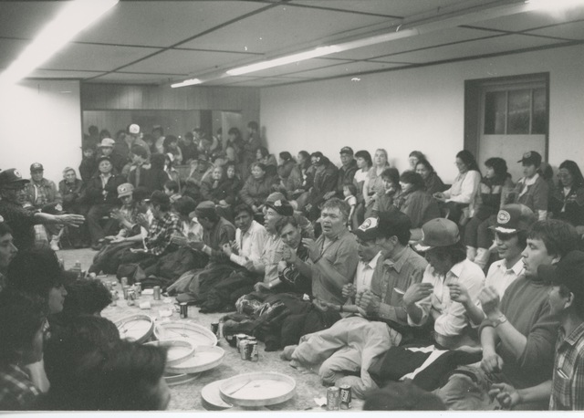 Large group gathered in a community hall, with men kneeling in a row and hand drums placed in front of them, while others watch.