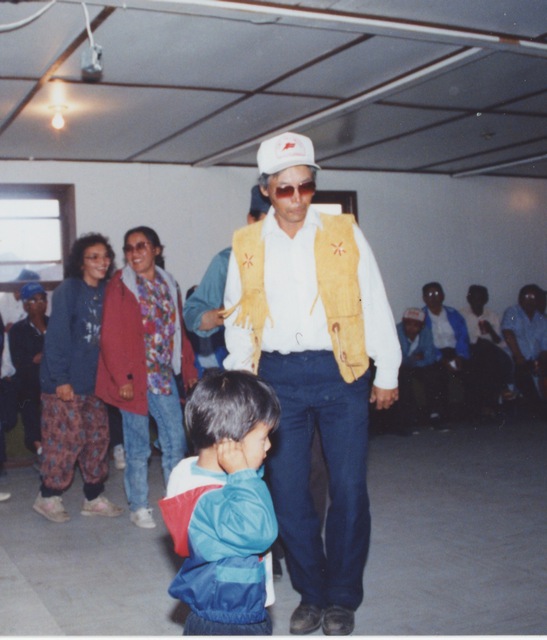 People gathered in a community hall, with Charlie Neyelle dancing in the centre wearing a fringed vest and cap, and a child standing in the foreground.
