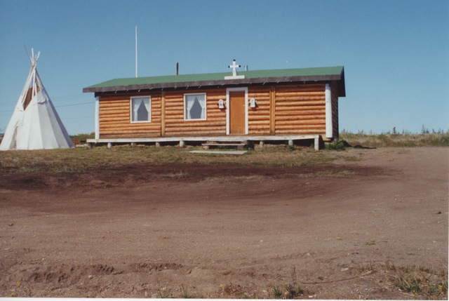 Log building with a cross above the entrance and a tipi nearby on a grassy clearing.