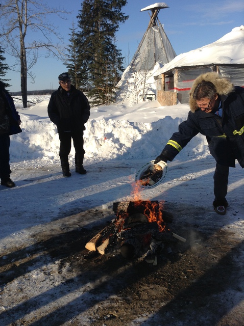 People gathered around a fire outdoors in winter, with Walter Bezha pouring an offering onto the flames.