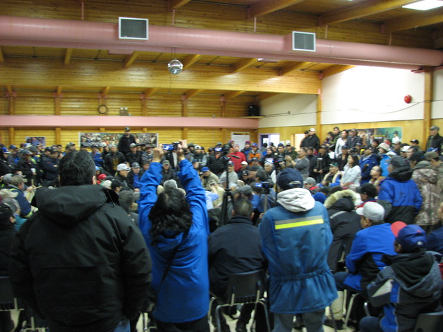Indoor gathering of adults and youth seated and standing, with some recording the event.