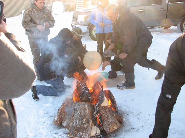 People gathered outdoors in winter around a fire, with one person holding a hand drum.