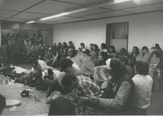 Large indoor gathering with a row of men kneeling and more men playing hand drums behind them, while others observe.