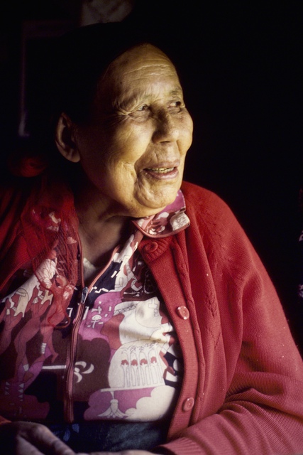 Louisa Baton seated and smiling, wearing a red sweater over a patterned shirt.