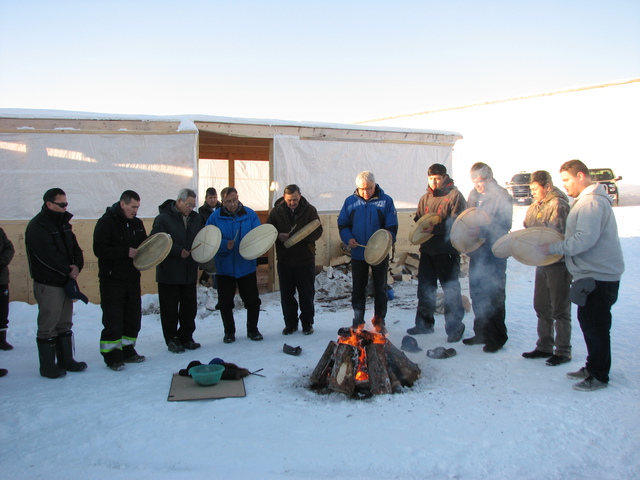 Men standing in a circle outdoors in the snow playing hand drums around a fire.