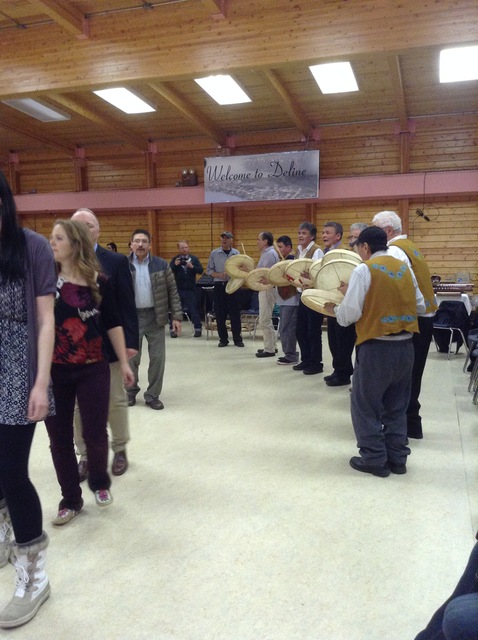 Men standing in a line playing hand drums and singing in a community hall, with a “Welcome to Délı̨nę” sign visible.