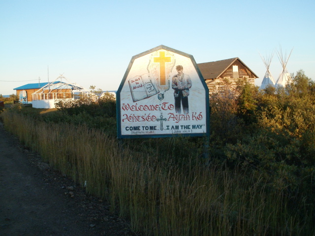 Hand-painted sign featuring a cross, a praying figure, and English and Dene text, with buildings and tipis in the background.