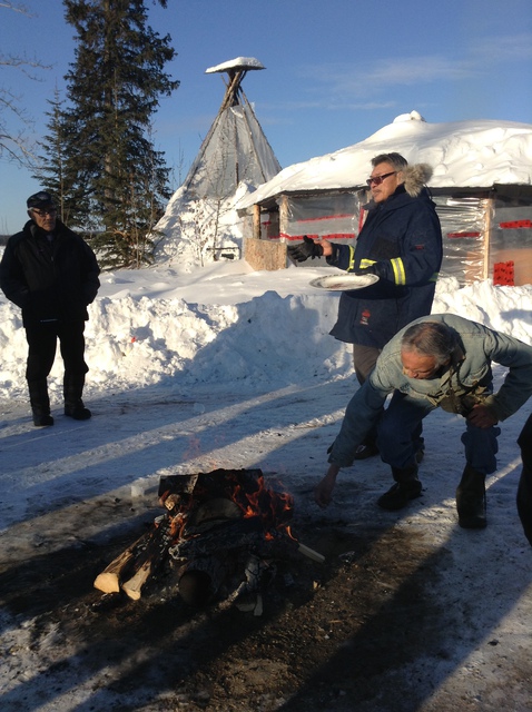 Walter Bezha and two other men standing by a small fire in a snowy landscape, with a building and tipi behind them.