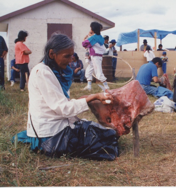 Jane Neyelle seated on grass scraping an animal hide, surrounded by community members during an outdoor gathering.