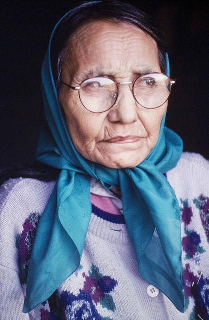 Portrait of Cecile Modeste wearing a teal headscarf, round eyeglasses, and a patterned knit sweater, seated against a dark background.