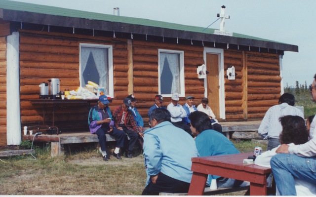 People seated outdoors in front of a log building with a cross above its entrance; a table set with refreshments is behind them.