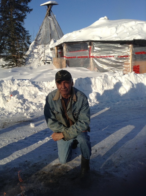 Michael Neyelle kneeling on a snow-covered road outside a building, with trees and a tipi in the background.