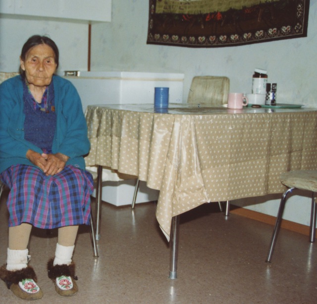 Bella Modeste seated beside a kitchen table, wearing a blue cardigan, a plaid dress, and moccasins with floral beadwork.