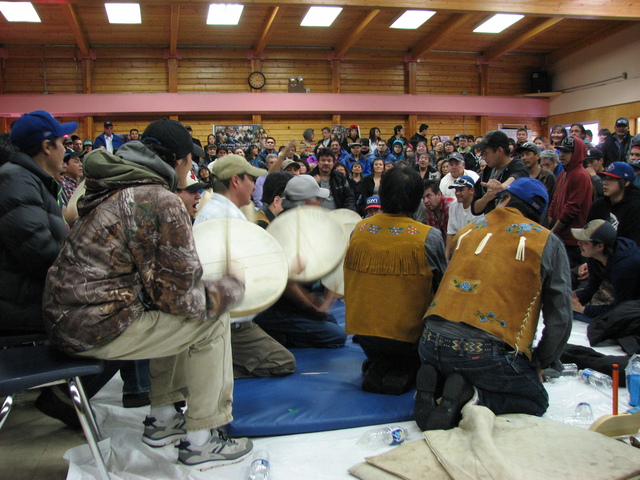 Men kneeling in a row with others behind them playing hand drums, surrounded by an audience.