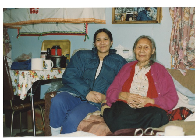 An elderly woman and a younger woman seated together in a domestic interior, surrounded by household items.