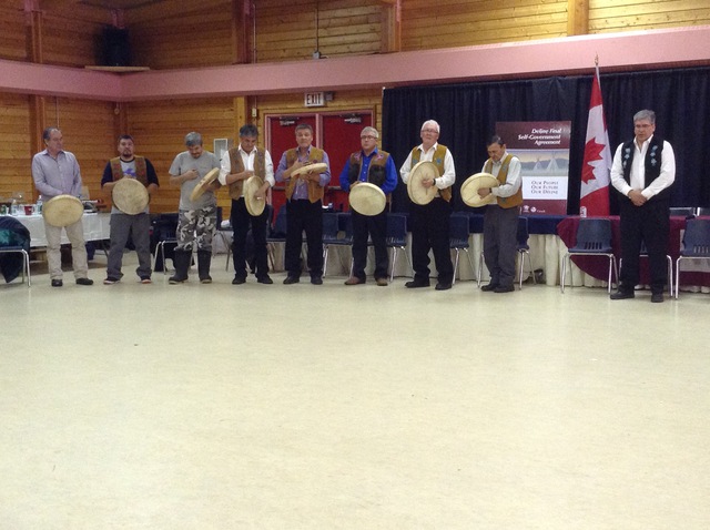 Men standing in a line holding hand drums in a community hall.