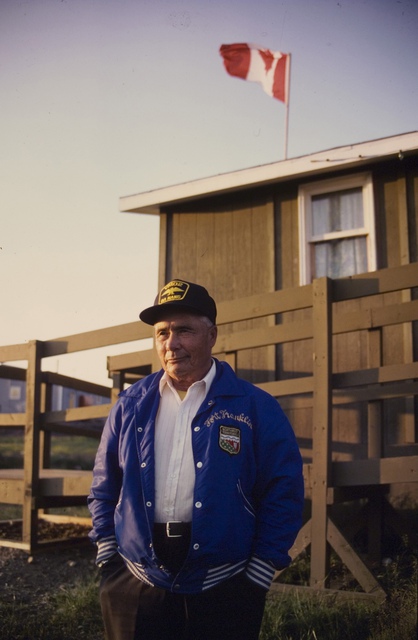 Paul Baton standing outdoors in front of a wooden building, wearing a blue jacket embroidered “Fort Franklin” and a cap, with a Canadian flag visible above the building.