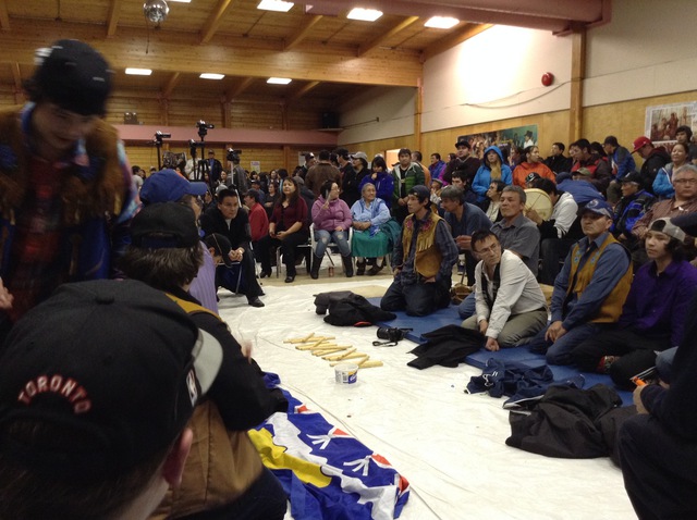 Participants kneeling on a mat indoors with sticks laid out in front; an audience surrounds them with cameras documenting the event.