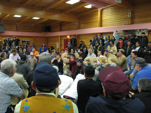 Men seated in a row playing hand drums and singing, with an audience observing.