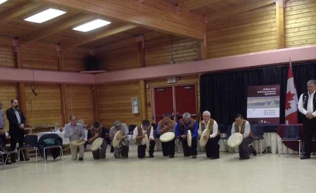 Men kneeling in a row in a community hall holding hand drums, with a Canadian flag and banner visible.