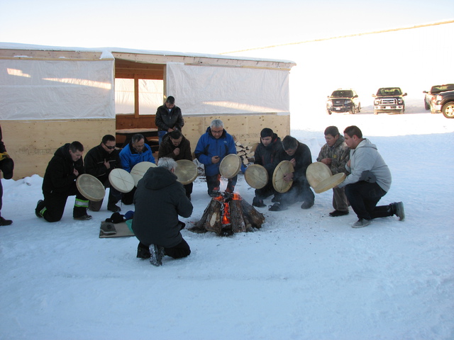 Men kneeling in a circle in the snow around a fire, holding hand drums, with a building and several parked vehicles nearby.